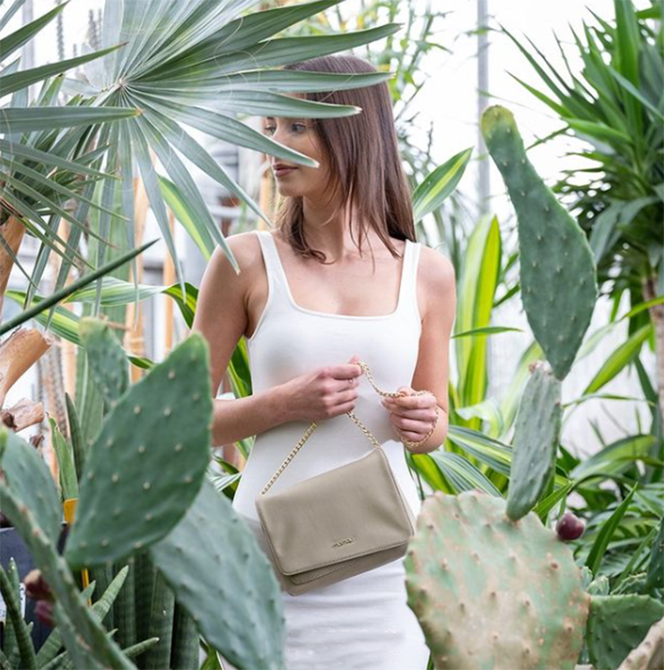 Ragazza con vestito bianco tra cactus e piante con sguardo rivolto a destra e in mano una borsetta vegan