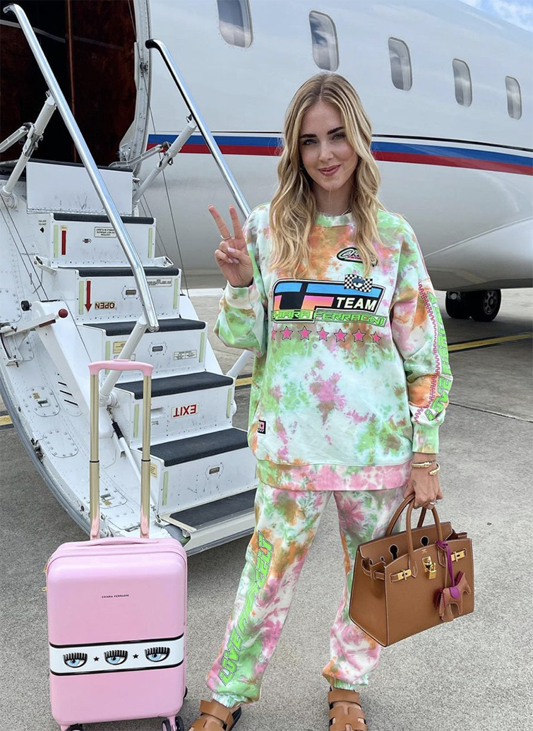 Chiara Ferragni davanti ad un aereo con la sua valigia e la sua borsa che fa segno con la mano destra